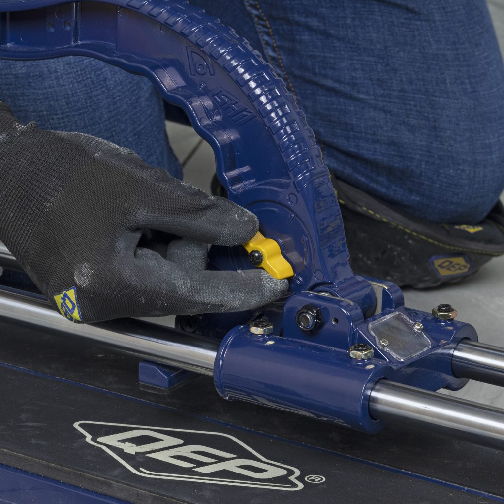 Installer making an adjustment to the height of the cutting wheel on the QEP 28" Big Clinker Tile Cutter while wearing QEP SureGrip Tiler's Gloves and ProMAX Gel Knee Pads